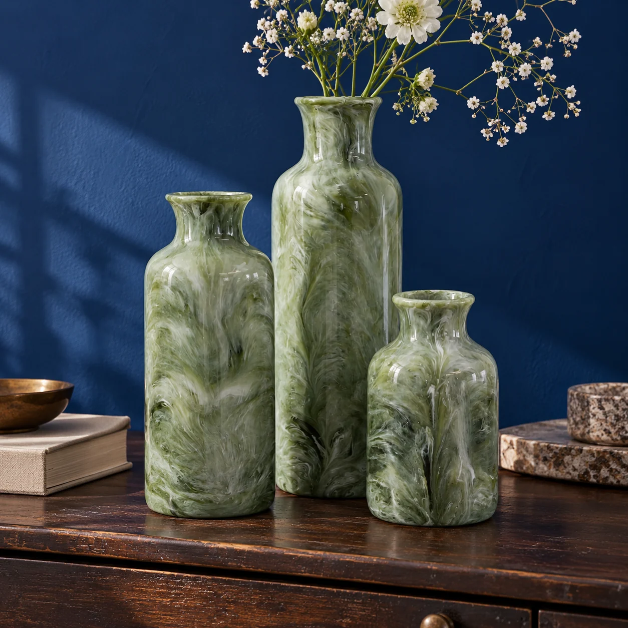 Three sage green marbled vases with white flowers on a dresser against a blue wall.